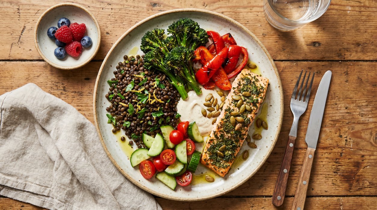 Balanced Mediterranean-style dinner plate with salmon, lentils, broccolini, roasted peppers, salad, olive oil, seeds, and berries.